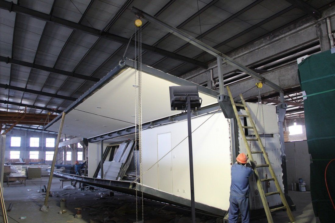 Worker near a two-story prefabricated building module inside a factory, suspended by a crane.
