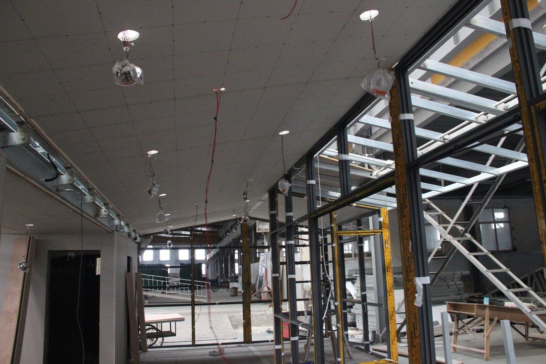 Interior construction site with exposed wiring and metal framing, recessed lights in ceiling, glass and metal walls.