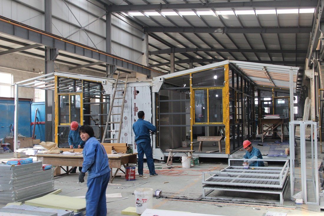 Factory interior: Workers assembling modular buildings with yellow window frames.