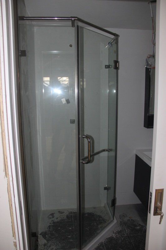 Glass shower stall with silver trim, open door. White walls, dark flooring, sink visible.