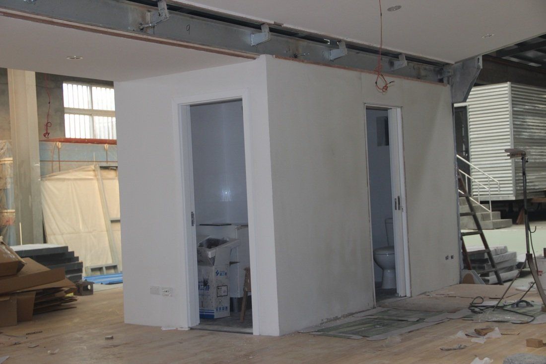 Two white-walled bathroom stalls under construction inside a building with visible beams and unfinished elements.