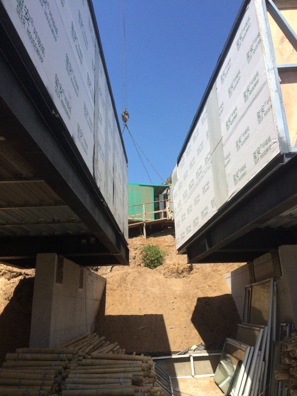 Two rectangular structures lifted by a crane over a hillside construction site with blue sky.