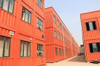 Red shipping container buildings stacked, forming multi-story structures with windows. Outdoors, bright day.