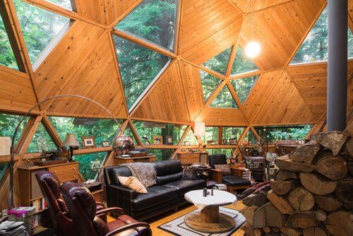 Cozy interior of a wood-paneled geodesic dome home with windows overlooking lush trees. Leather furniture and a wood-burning stove are present.