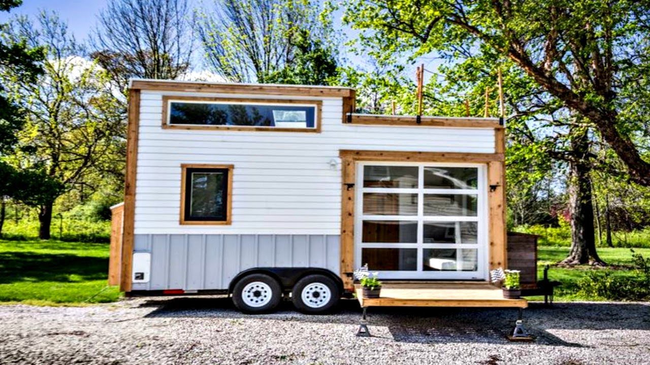 Tiny white house on wheels with a glass garage door and wooden accents in a grassy setting.