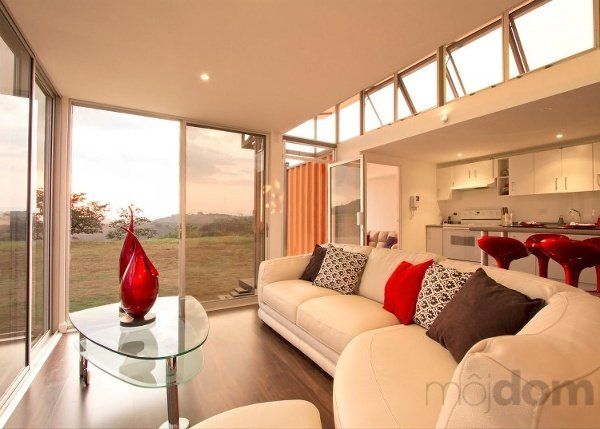 Modern living room with white sofa, glass table, and large windows overlooking a field.