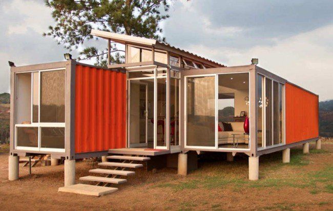 Orange container home with glass walls, steps, and elevated on concrete pillars.