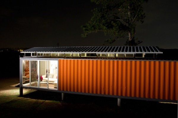 Orange shipping container house at night, lit interior, solar panels on roof.