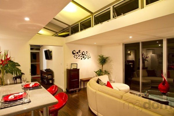 Modern living room with white walls, tan curved sofa, and a kitchen bar with red chairs.