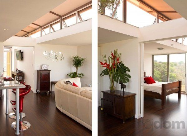 Modern interior with white walls, dark wood floor, and large windows.  Red bar stools, sofa, and bedroom with wooden bed frame.