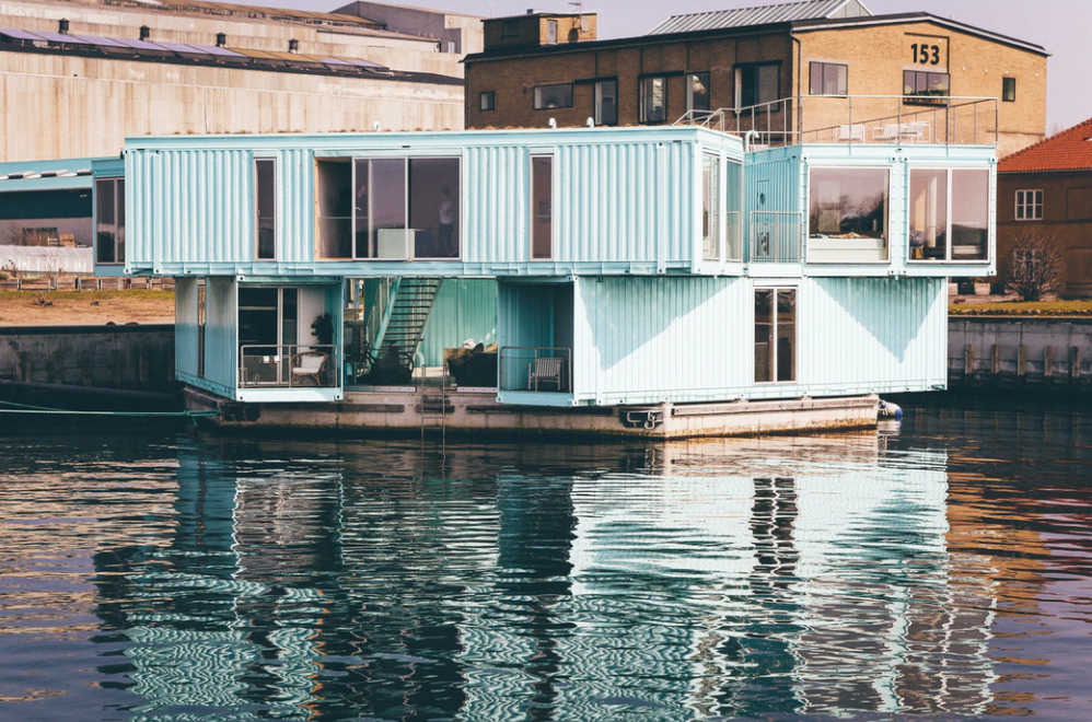 Aqua-colored shipping container home floating on water, with reflections in the ripples.