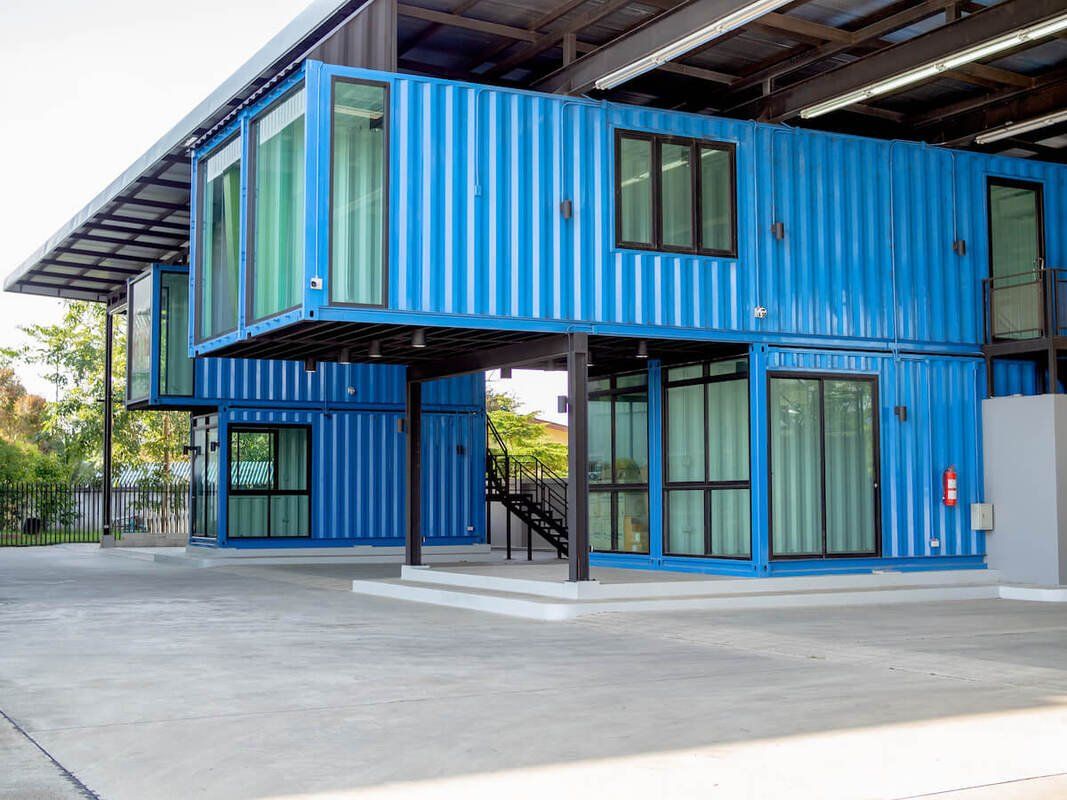 Blue shipping container home with windows, elevated on black supports, under a metal roof.