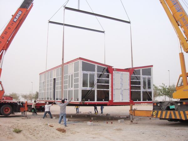 Two cranes lifting a modular building with multiple windows. Workers are guiding the structure at a construction site.