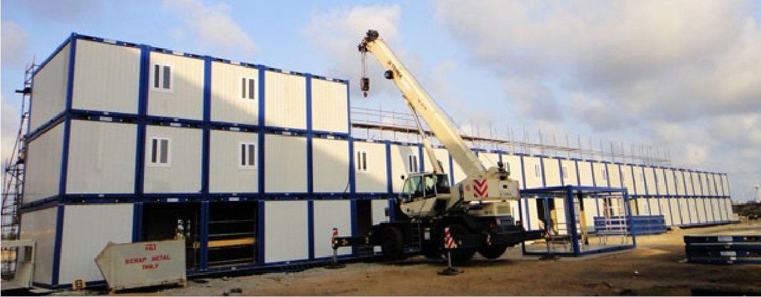 Construction site with modular buildings being assembled by a crane under a cloudy sky.