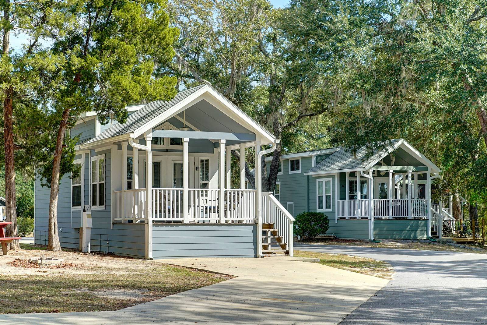 Two light blue cottage-style houses with white porches and stairs, set in a grassy area with trees.
