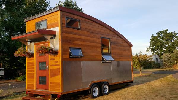 Tiny home trailer with wooden siding and metal lower section, steps, flowers, and a curved roof.