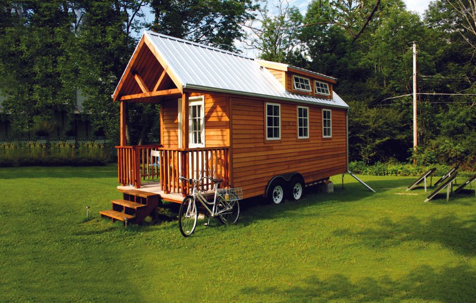 Tiny wooden house on wheels with a porch, parked on a green lawn, bicycle nearby.