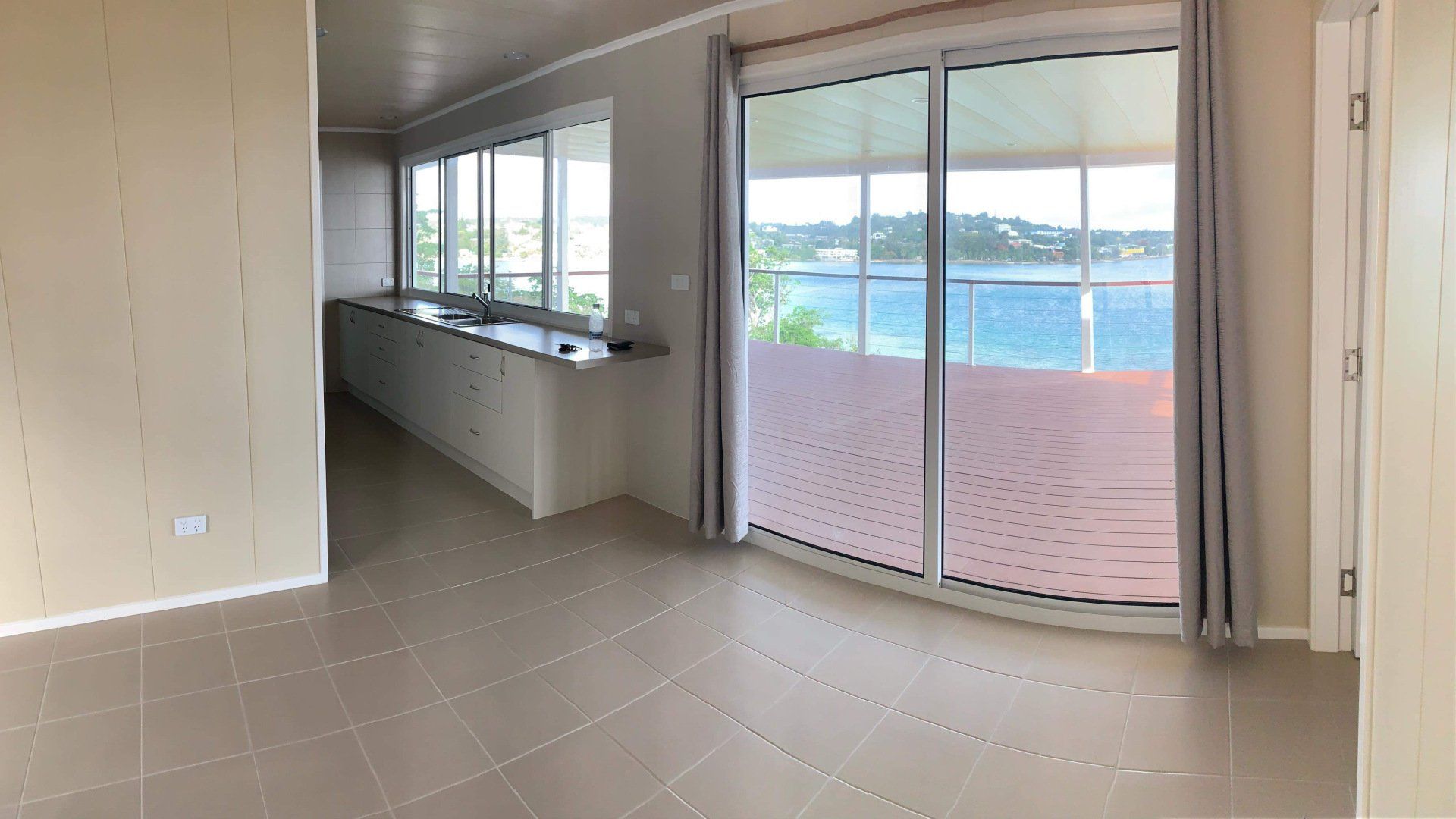 Empty living area with large windows overlooking a blue ocean. Beige tiled floor, light walls, and sliding glass door.