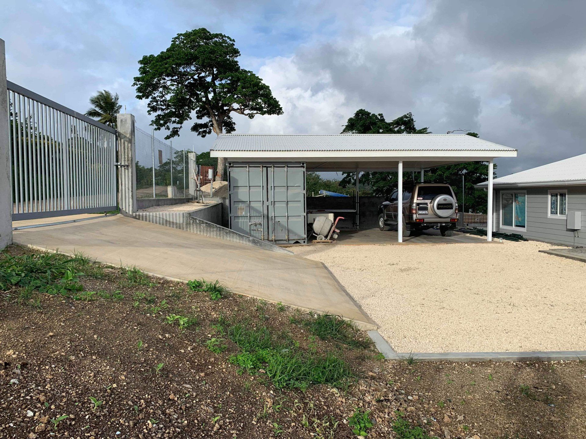 A gravel driveway leads to a carport with a vehicle. A concrete ramp and fence are on the left.