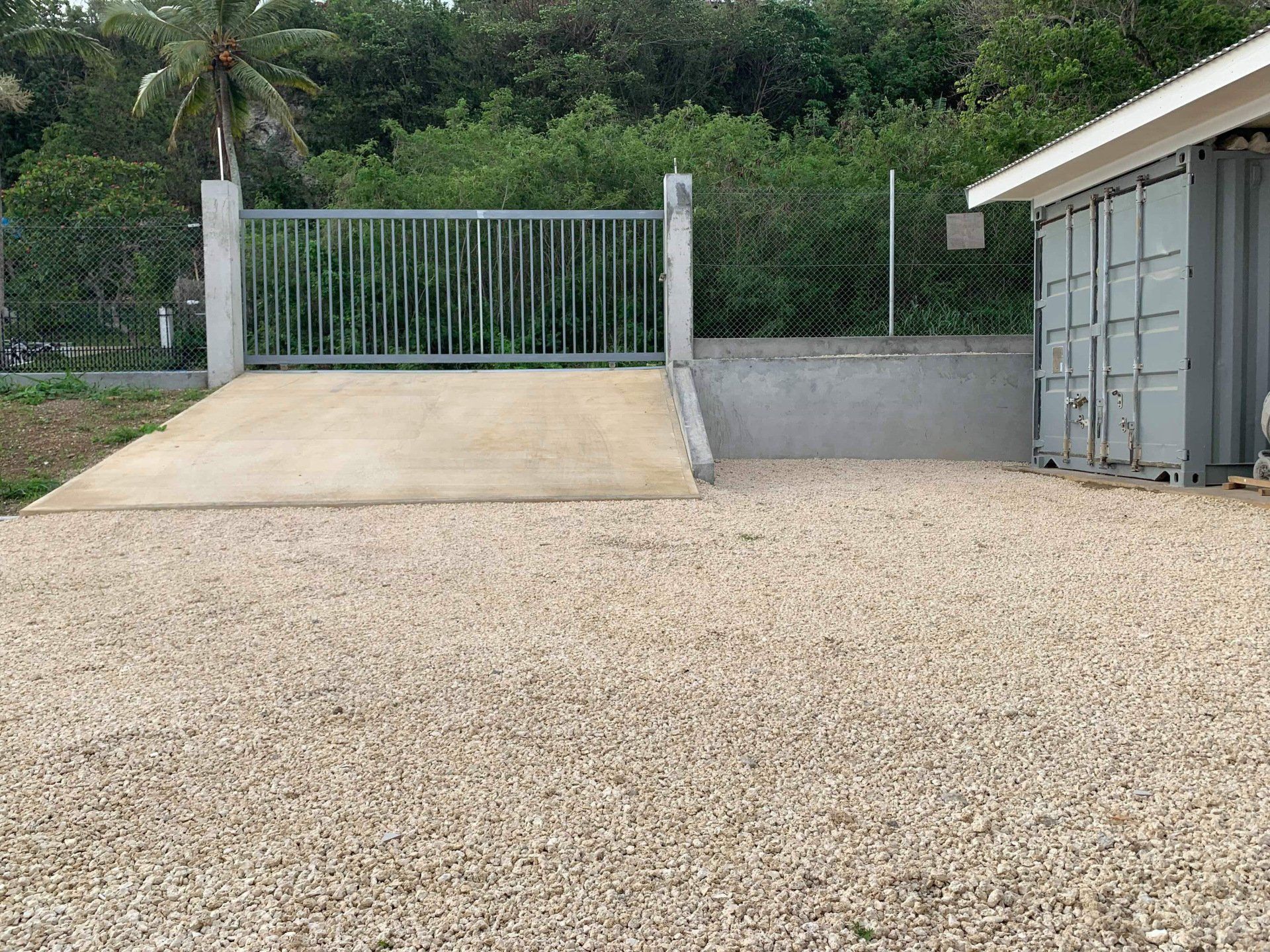 Gravel lot with concrete ramp leading to a metal gate. Two shipping containers sit to the right, trees in the background.