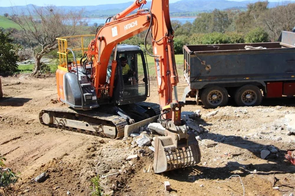 A Large Orange Excavator Is Sitting in A Dirt Field Next to A Dump Truck — Albury Demolitions in Lavington, NSW