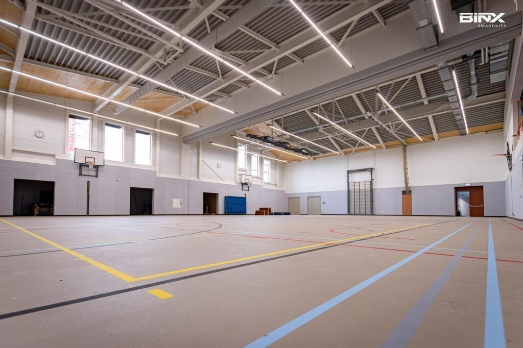 Een lege overdekte sporthal met een lichtgekleurde vloer, veelkleurige belijning en felle plafondverlichting.