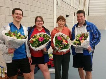 Vier mensen staan ​​in een gymzaal, elk met een glimlach op hun gezicht en een groot boeket bloemen in hun handen