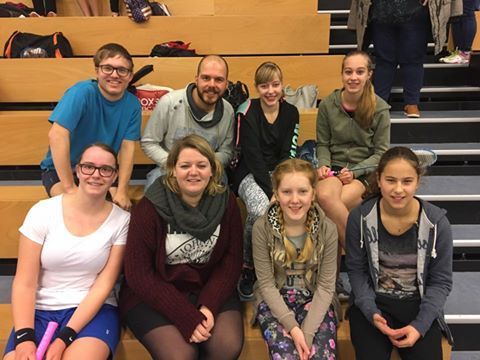 Een groep van acht mensen zit lachend op houten tribunes in een overdekte sporthal.