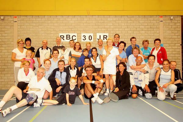Een grote groep poseert op een badmintonveld voor een muurbord met de tekst 