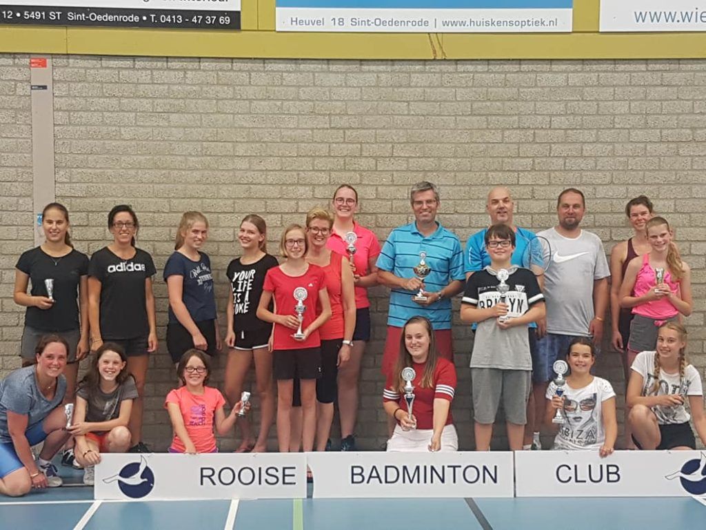 Een groep mensen staat in een gymzaal te poseren met trofeeën voor een bord van de Rooise Badminton Club.