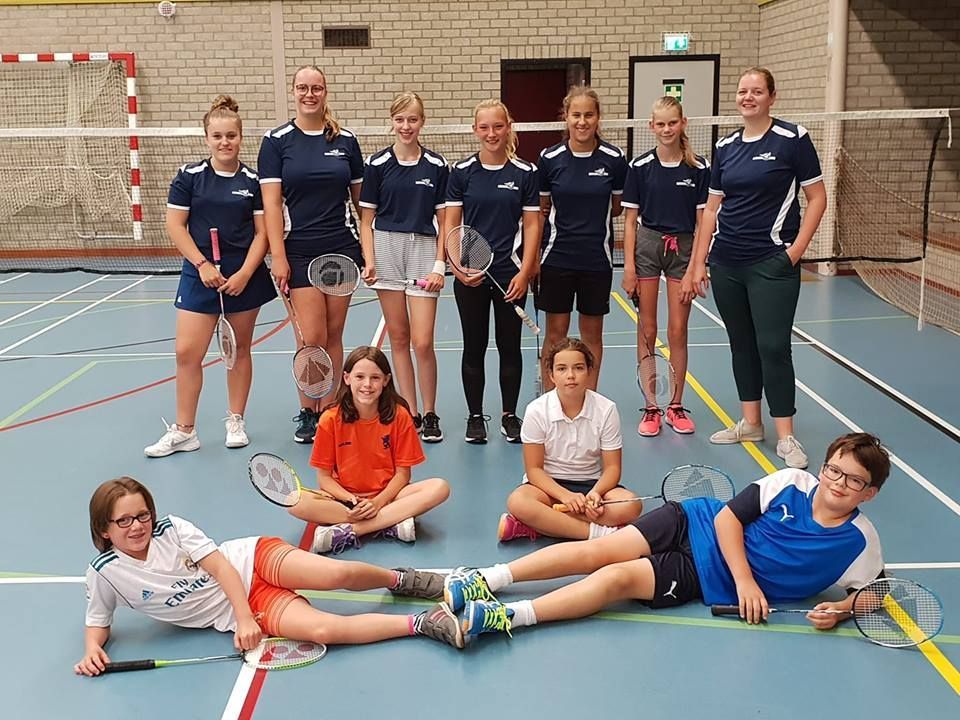 Een groep mensen in sportkleding poseert met badmintonrackets in een sportschool. Ze staan ​​en zitten op een blauwe baan.