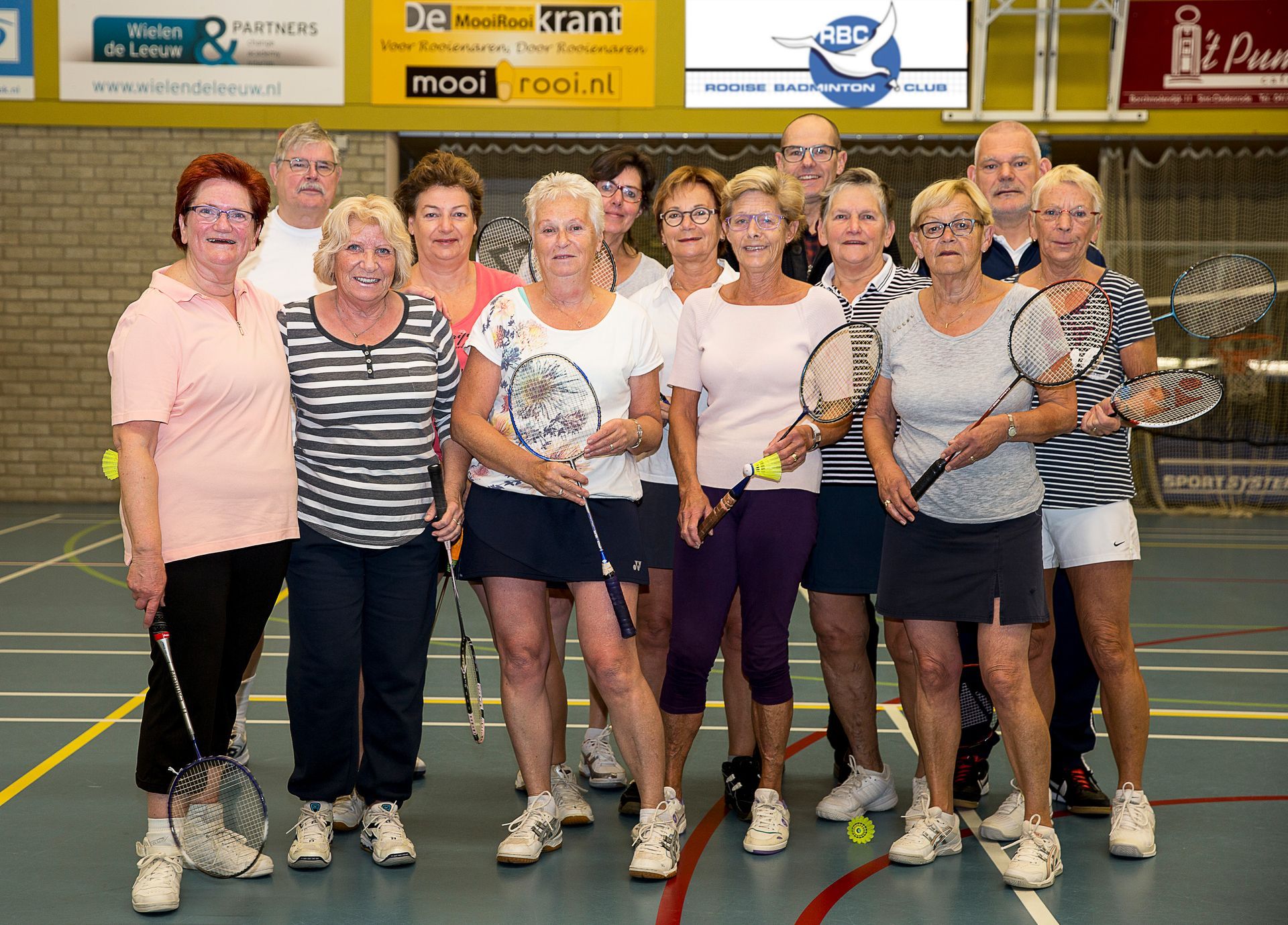 Een groep mensen staat samen in een sporthal met badmintonrackets in de hand voor een groepsfoto.