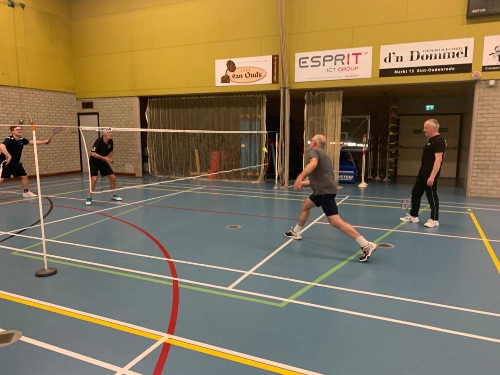 Vier mensen spelen badminton op een binnenbaan met een blauwe vloer en felgele muren.