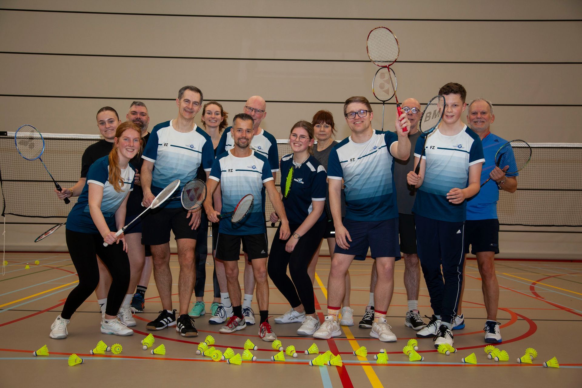 Een groep badmintonspelers in sportkleding poseert met rackets in een sporthal, terwijl de shuttlecocks op de grond liggen.