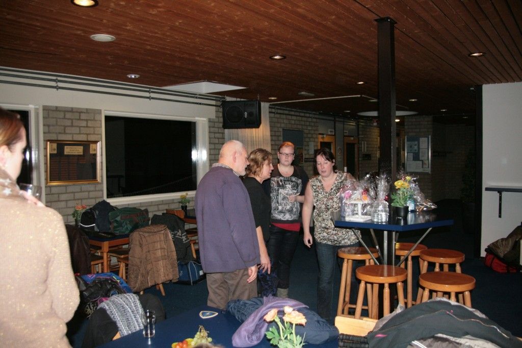 Een groep mensen staat met cadeaus rond een tafel in een met houten panelen beklede binnenruimte.
