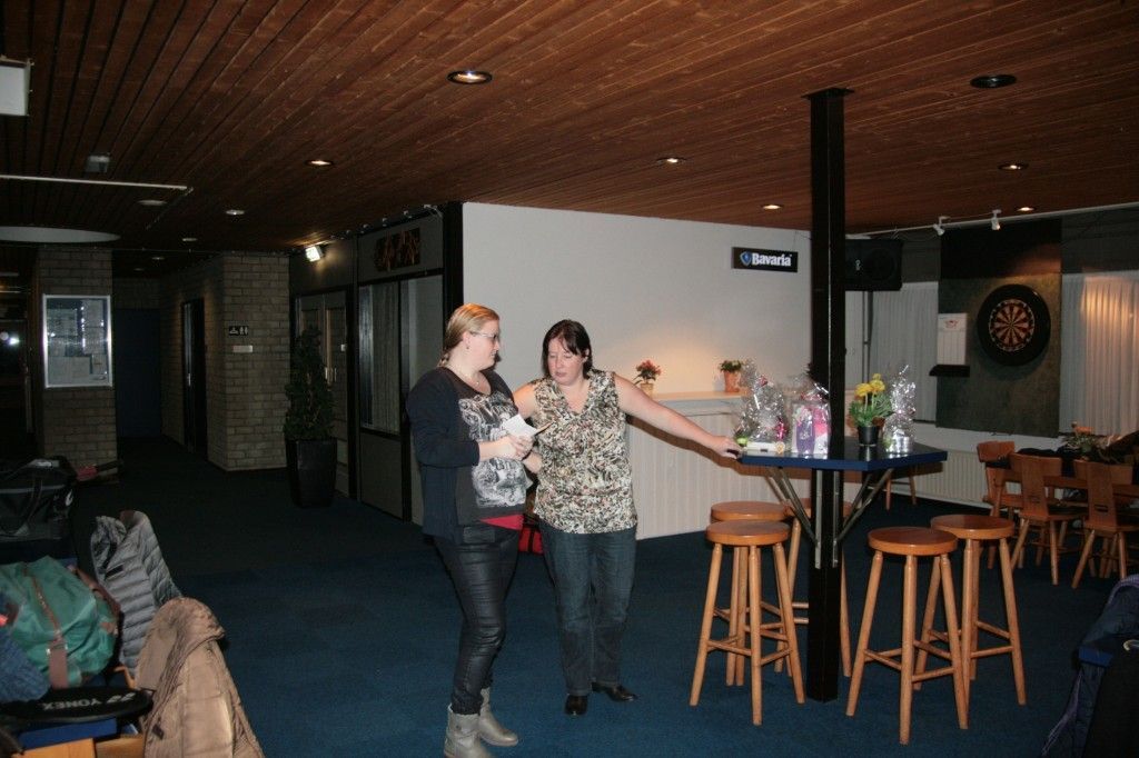 Twee mensen staan ​​te praten in een schemerig verlichte lounge met een dartbord, houten barkrukken