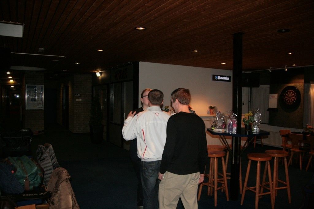Drie mensen staan ​​bij elkaar in een schemerig verlichte bar en voeren een gesprek nabij een hoge tafel met houten krukken.