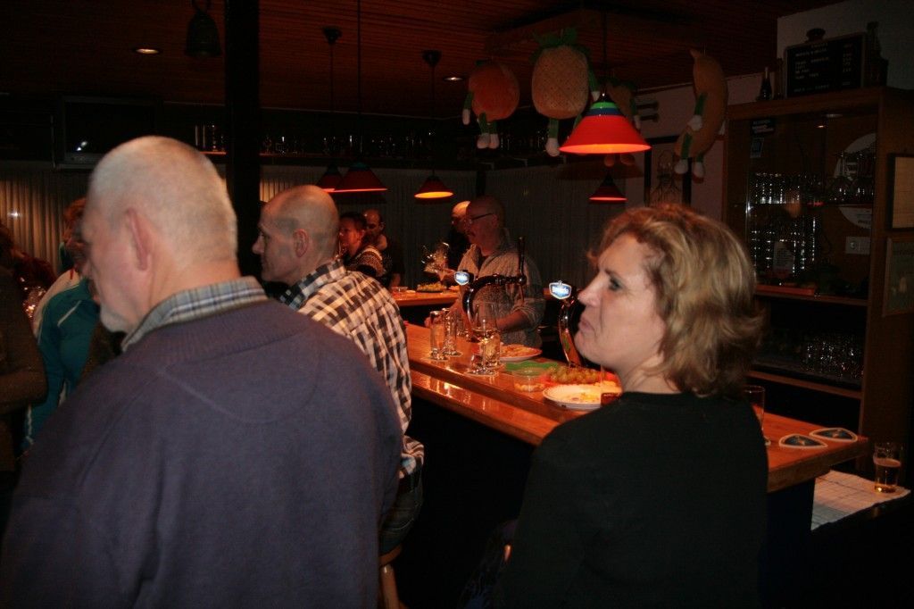 Mensen verzamelen zich aan een schemerig verlichte bar, waar ze tegenover elkaar aan de bar zitten onder hangende lampen