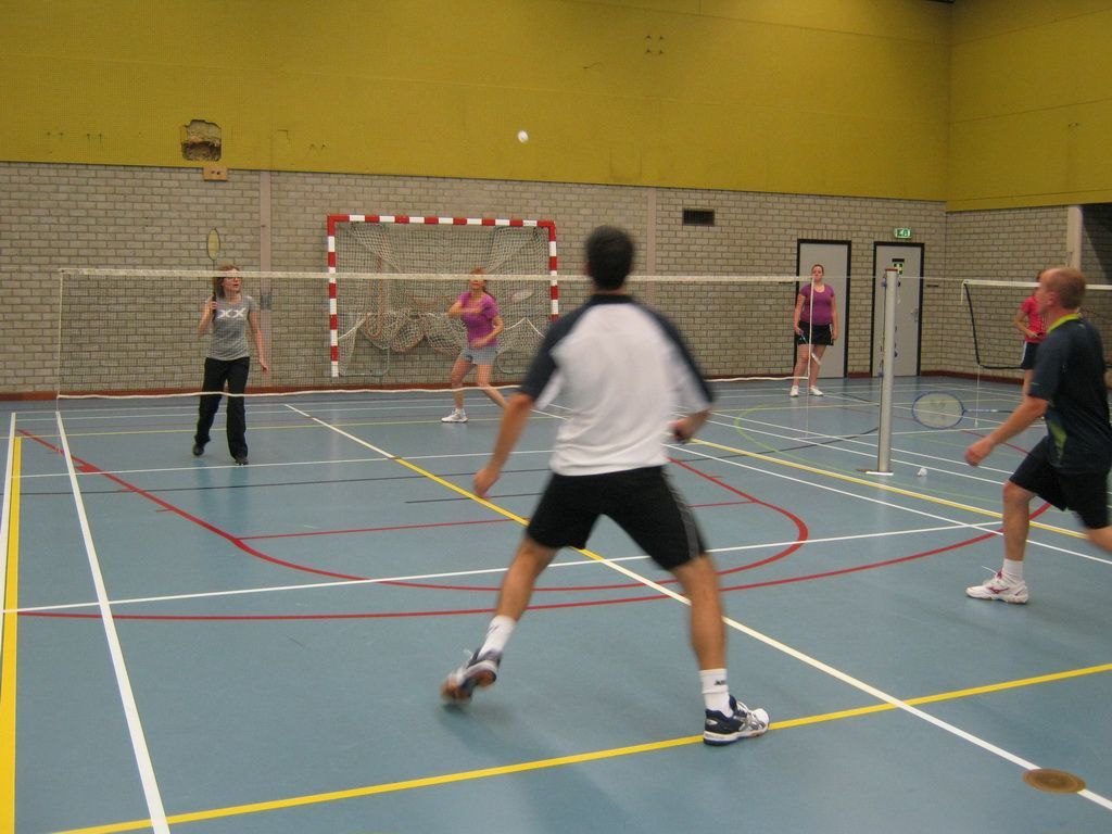 Vier mensen spelen badminton in een binnengymzaal met een blauw veld, een net en een handbaldoel op de achtergrond.