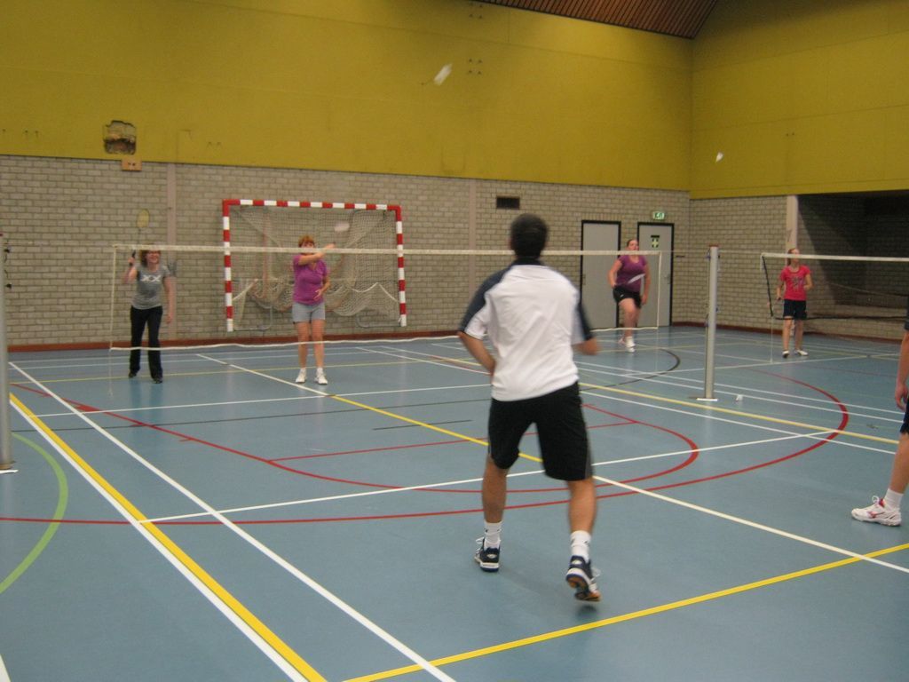 Een groep mensen speelt badminton in een overdekte sporthal met een blauwe vloer en gele muren.