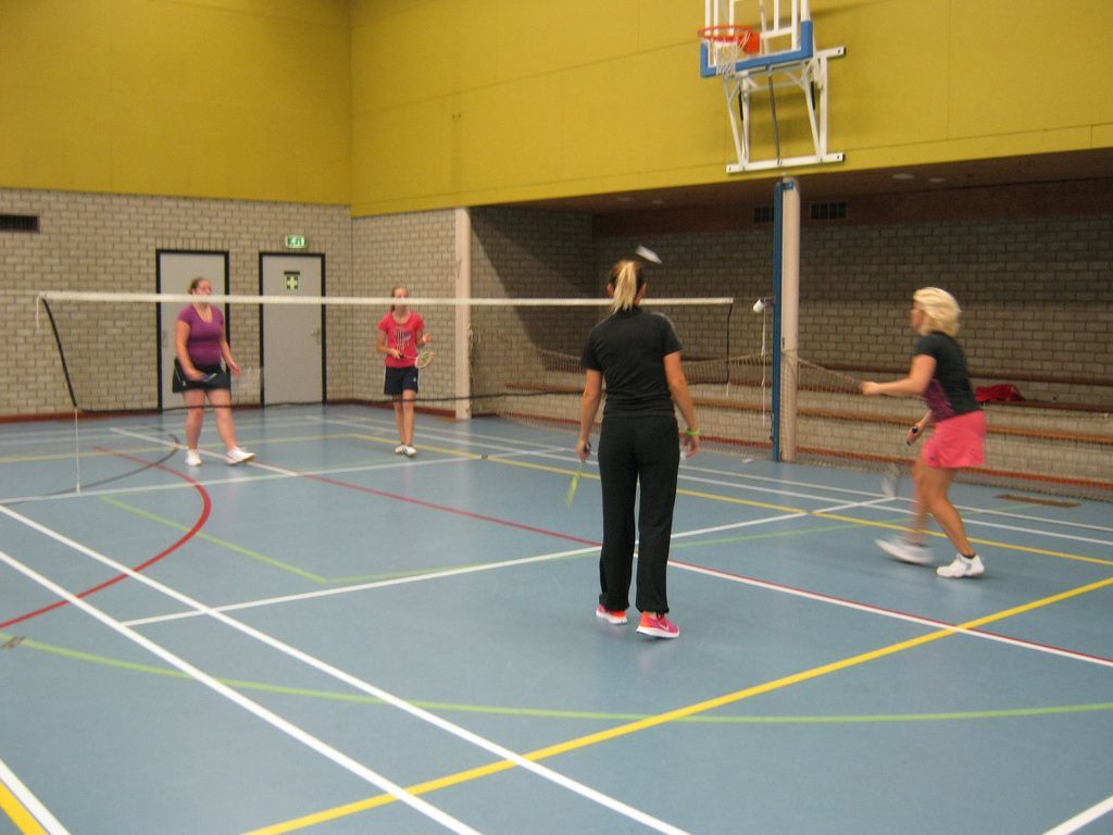 Vier mensen spelen badminton in een binnengymzaal op een lichtblauwe baan met witte lijnen.