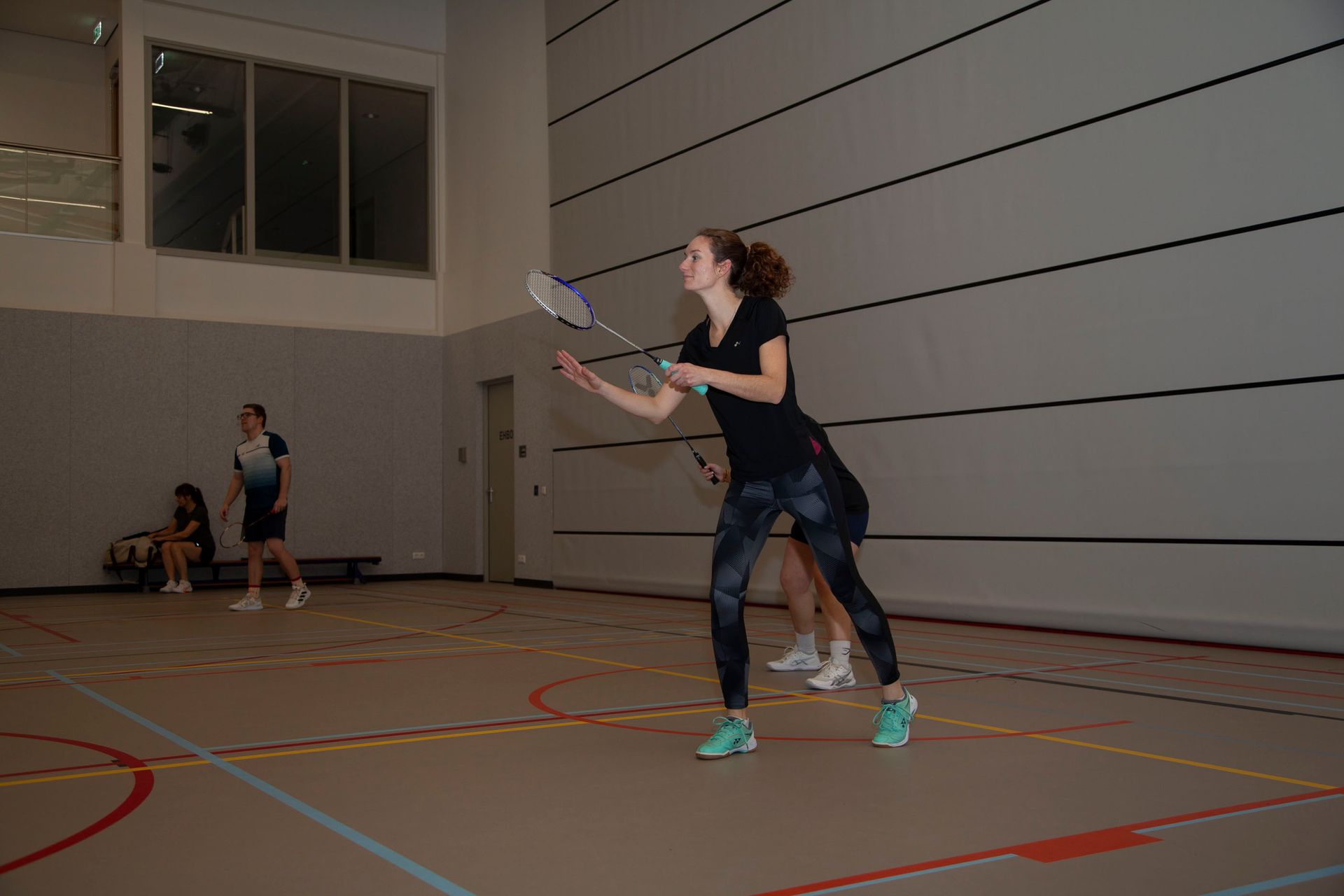 Een persoon in sportkleding houdt een badmintonracket vast in een voorwaartse uitvalspositie op een indoorbaan
