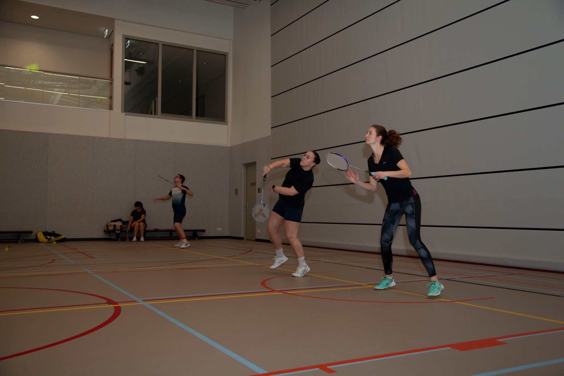Drie mensen spelen badminton in een binnengymzaal.