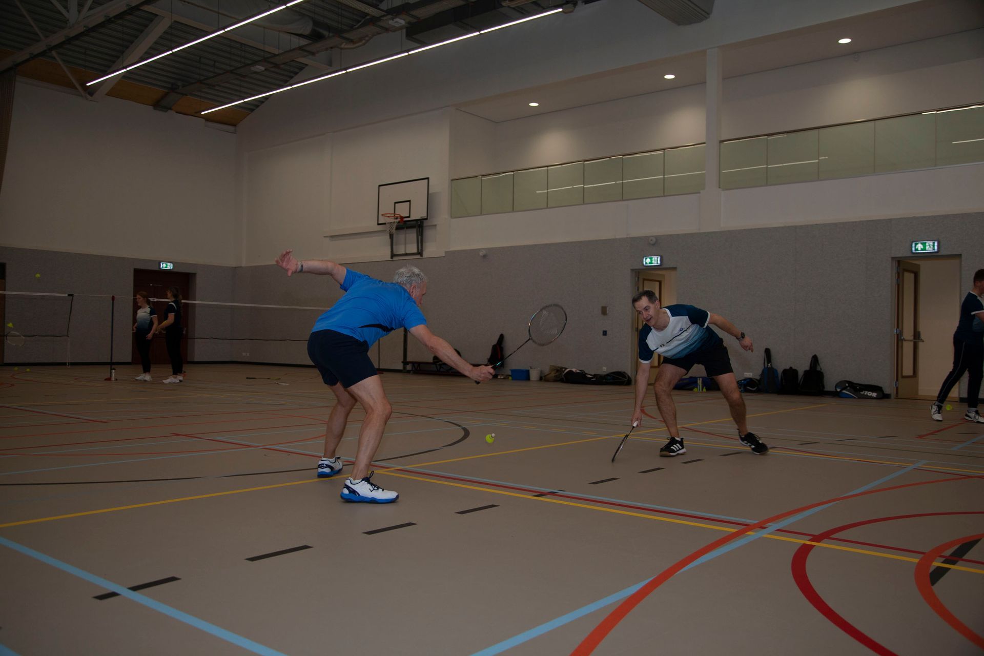 Twee mensen spelen badminton op een indoorbaan; de ene reikt naar een lage bal