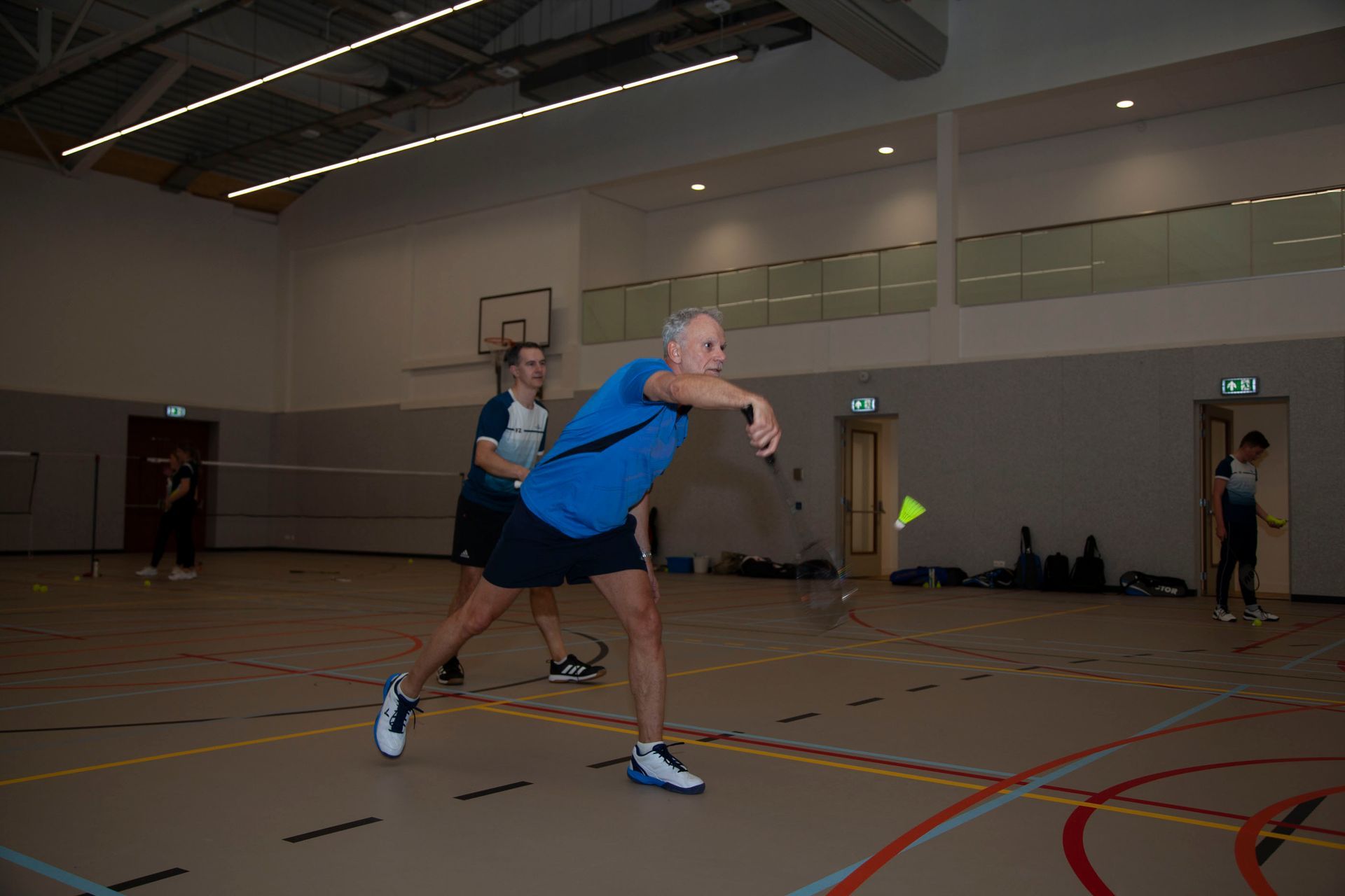 Een badmintonspeler slaat een shuttlecock in een sporthal, terwijl een andere speler achter hem op de baan staat.