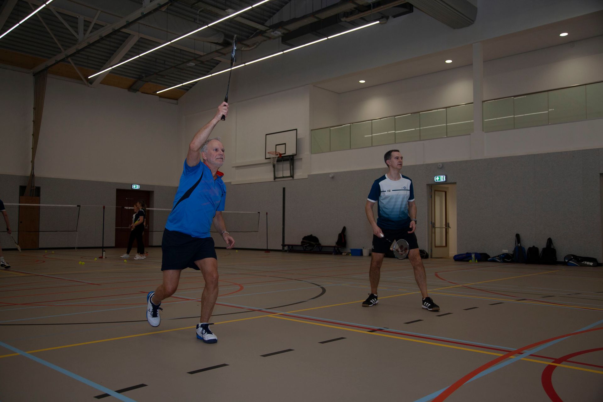 Twee mensen spelen badminton op een indoorbaan; de ene maakt zich klaar om de shuttle te slaan, terwijl de andere klaarstaat.