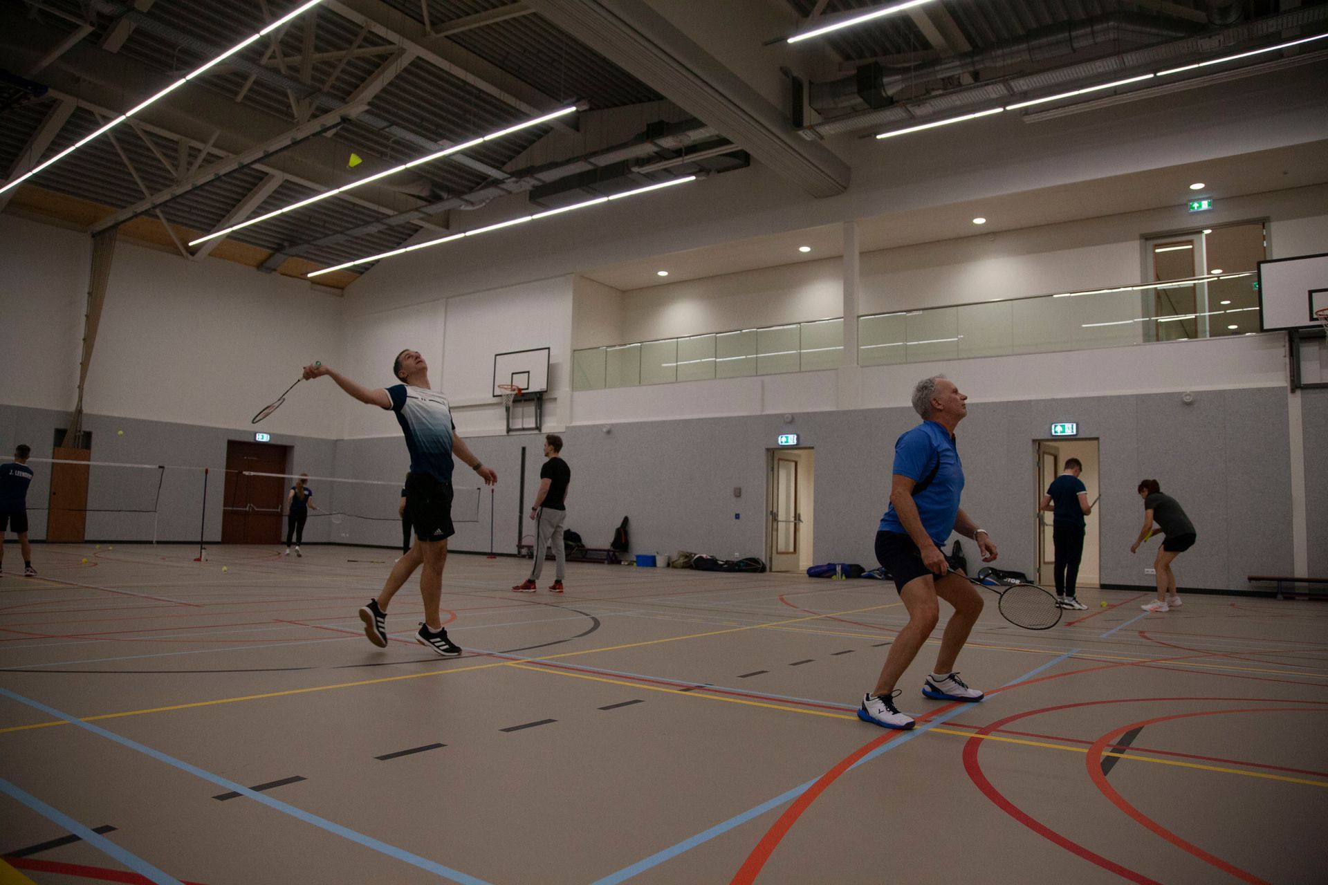 Mensen spelen badminton in een felverlichte overdekte sporthal met houten vloeren en veldmarkeringen.