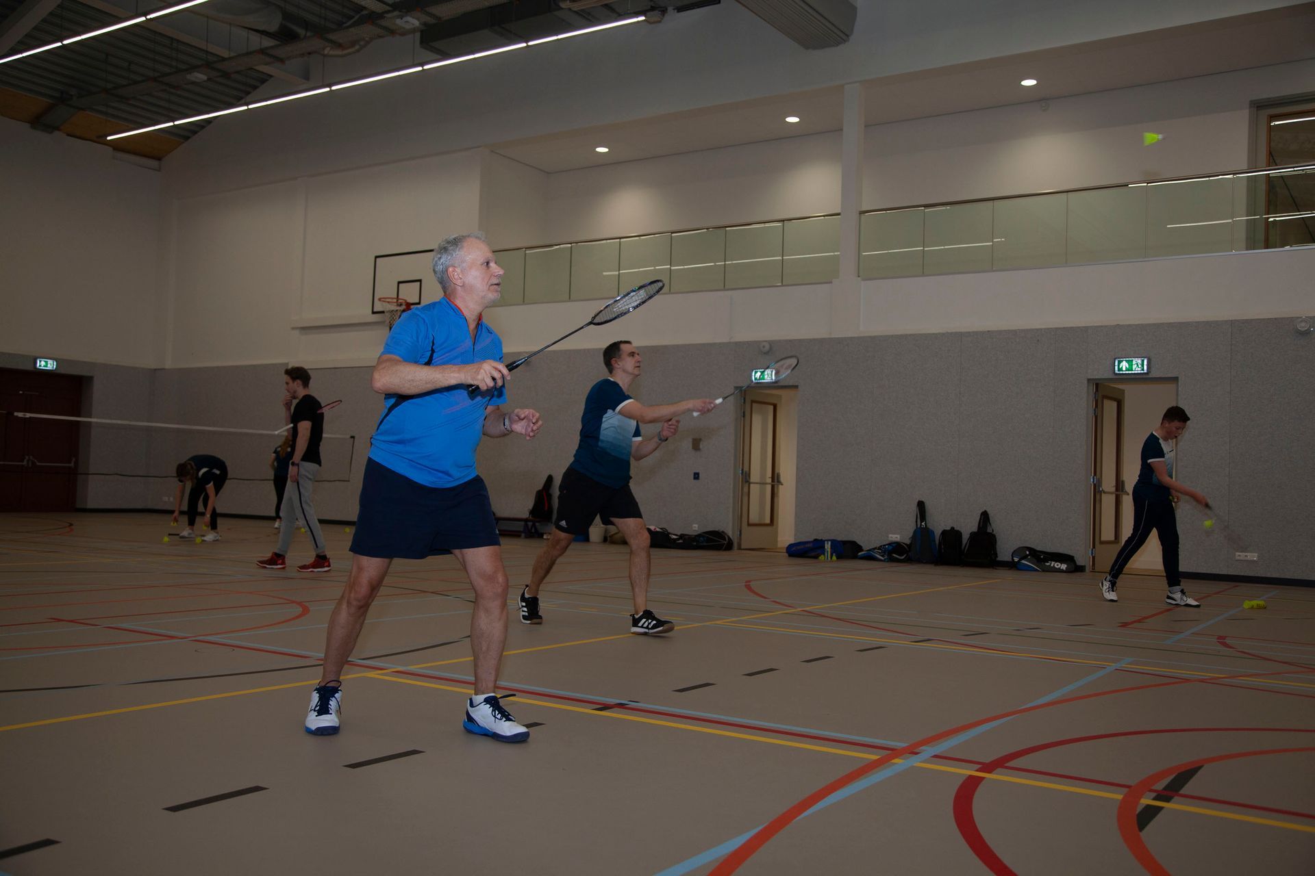 Een groep speelt badminton in een goed verlichte overdekte sporthal met houten vloeren en geschilderde belijning.
