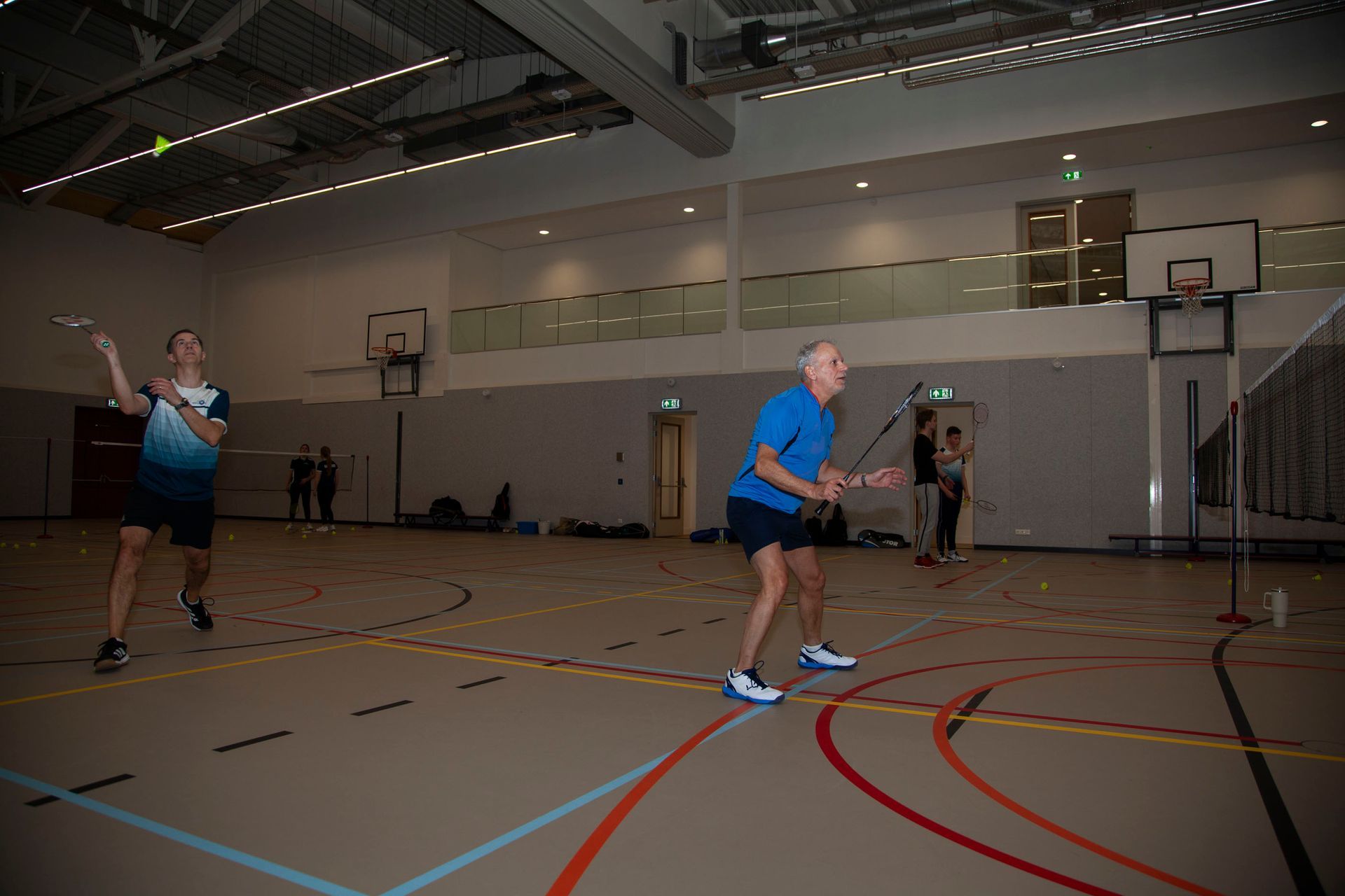 Twee mensen spelen badminton op een indoorbaan, beiden met een racket in de hand en geconcentreerd op het spel.