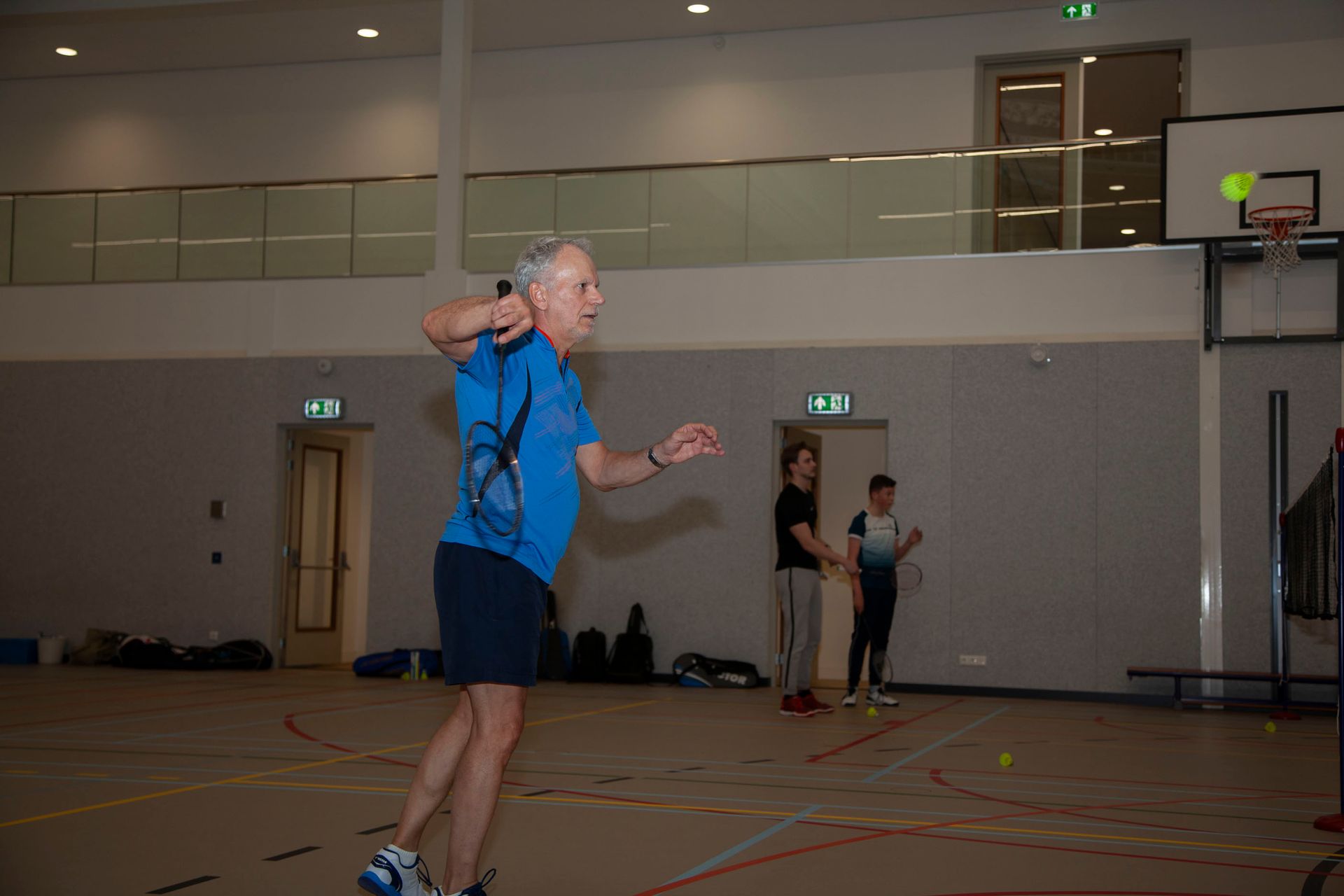 Een persoon in een blauw shirt houdt een badmintonracket vast in een overdekte sporthal.